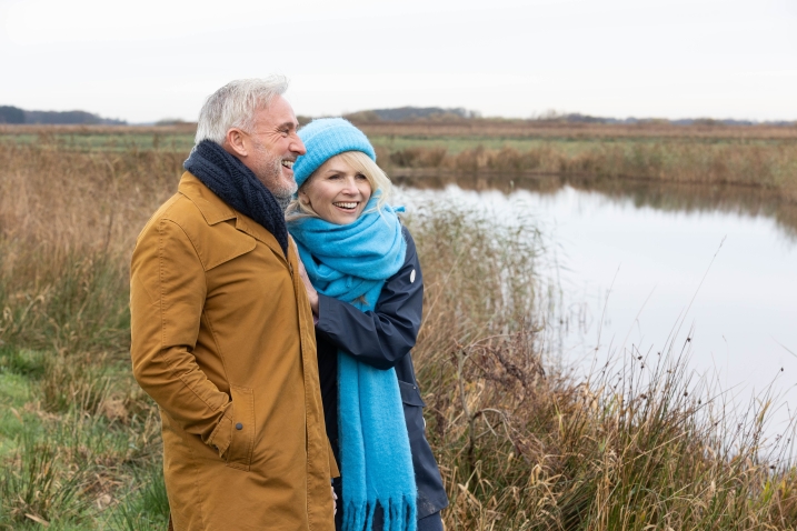 A couple, dressed for winter, walking beside water in a grassy wetland landscape. 